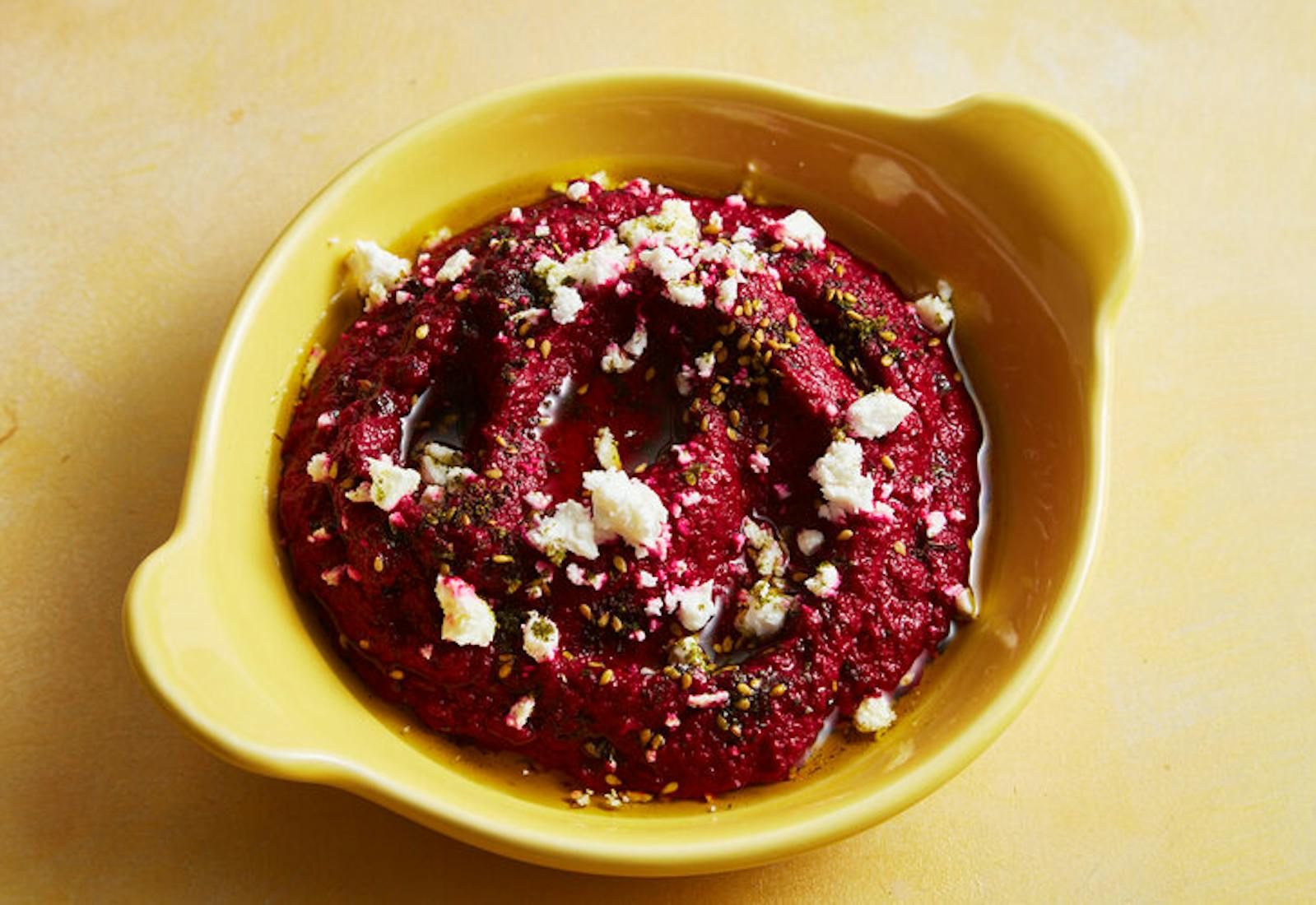 Spiced beet dip with crumbled feta in yellow dish atop yellow surface.