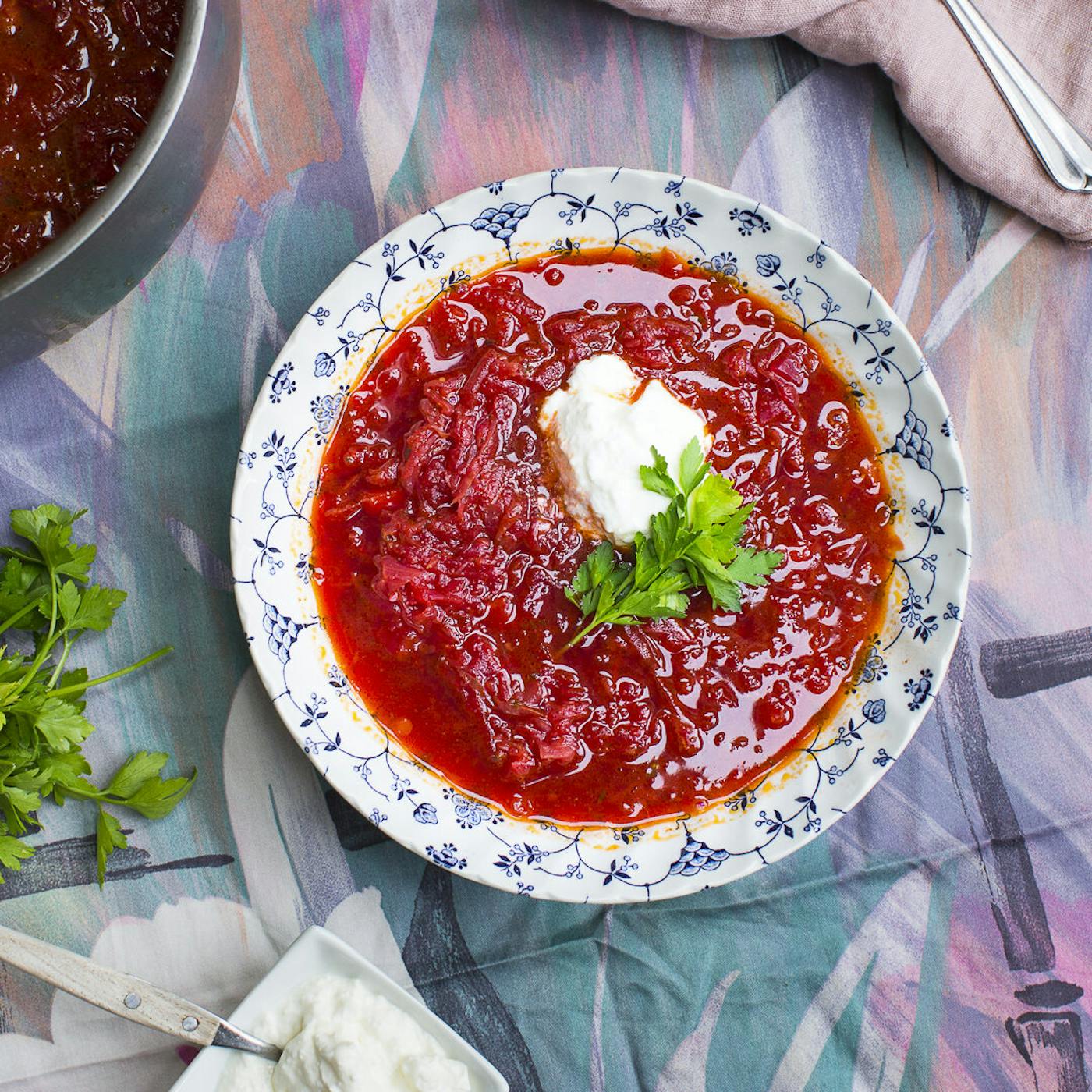 Borscht with dollop of sour cream and parsley alongside vegetarian borscht atop colorful tablecloth.