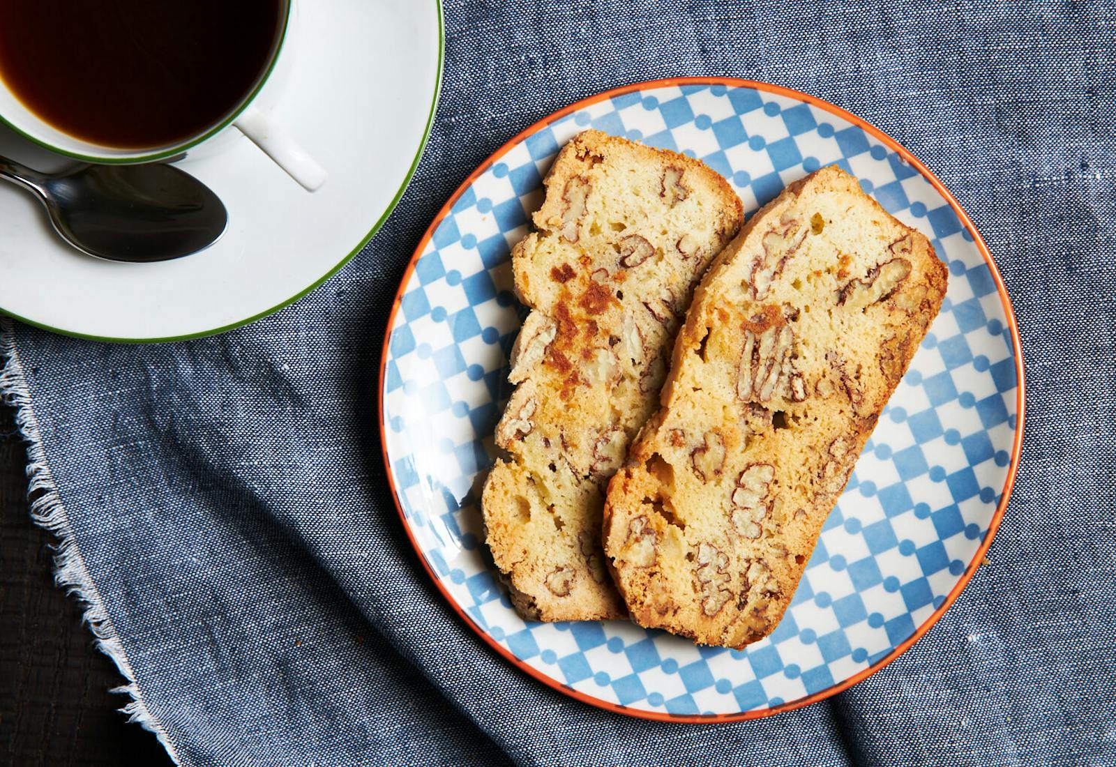 Slices of pecan mandel bread on blue and orange patterned dish with cup of coffee atop grey cloth.