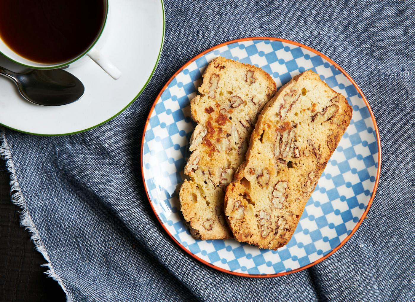 Slices of pecan mandel bread on blue and orange patterned dish with cup of coffee atop grey cloth.