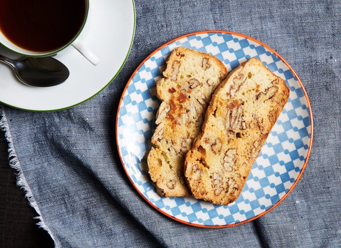 Slices of pecan mandel bread on blue and orange patterned dish with cup of coffee atop grey cloth.