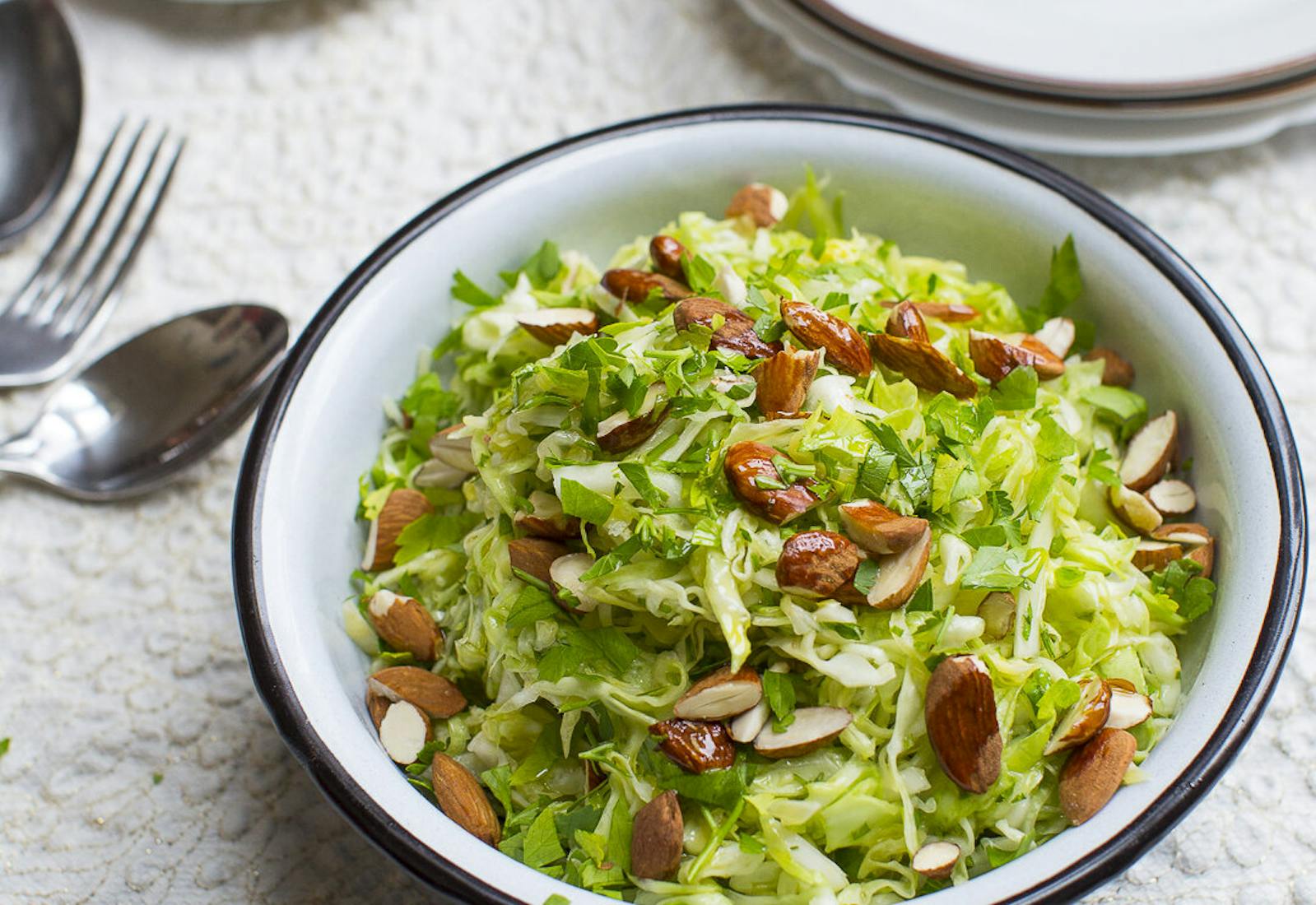 Cabbage salad with chopped almonds alongside dish with lemon wedges atop white tablecloth.