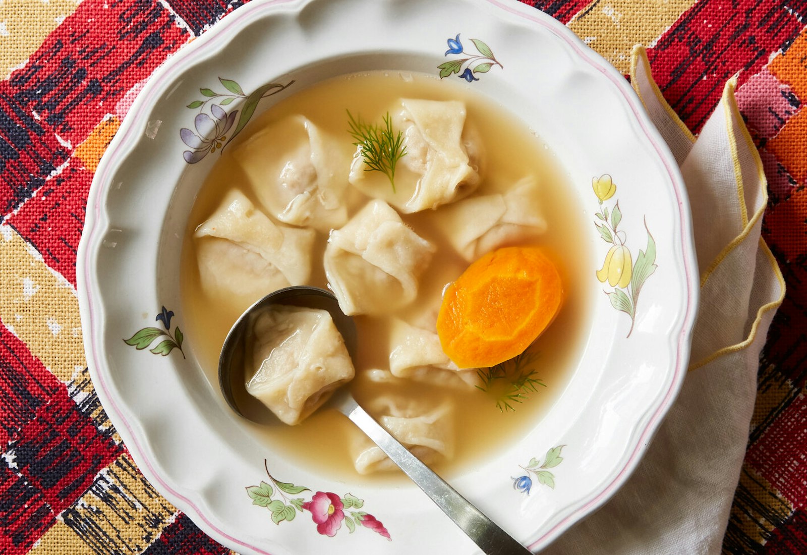 Chicken and liver kreplach with fresh dill in floral bowl atop red and yellow patterned tablecloth.