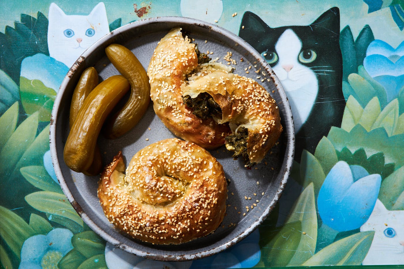 Bourekas with Israeli pickles on grey plate atop tablecloth with graphic cats and flowers.