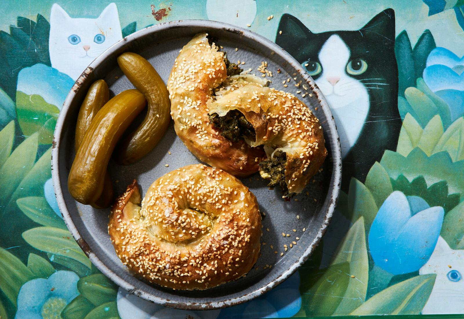 Bourekas with Israeli pickles on grey plate atop tablecloth with graphic cats and flowers.