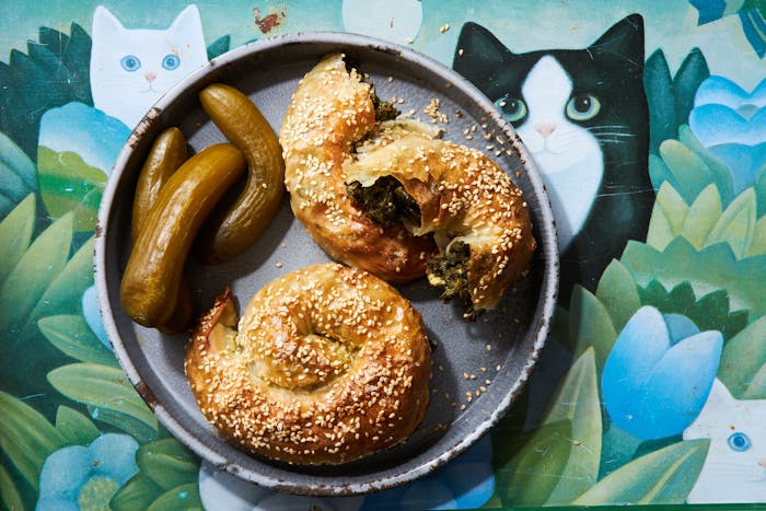 Bourekas with Israeli pickles on grey plate atop tablecloth with graphic cats and flowers.