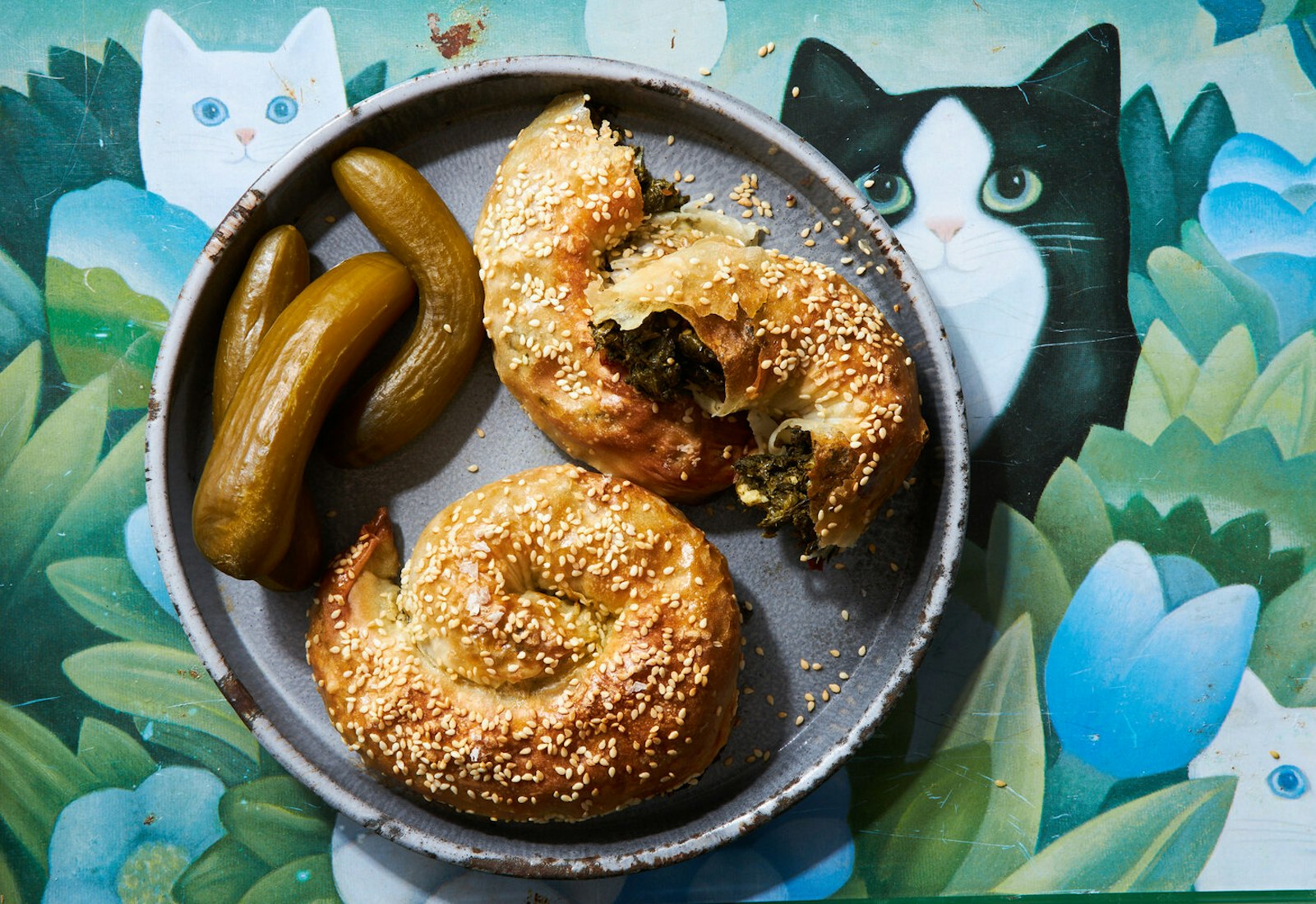 Bourekas with Israeli pickles on grey plate atop tablecloth with graphic cats and flowers.