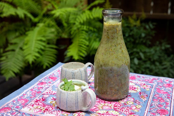 Glass bottle with schav alongside mugs of schav with sour cream and fresh sorrel atop printed tablecloth, outdoors.