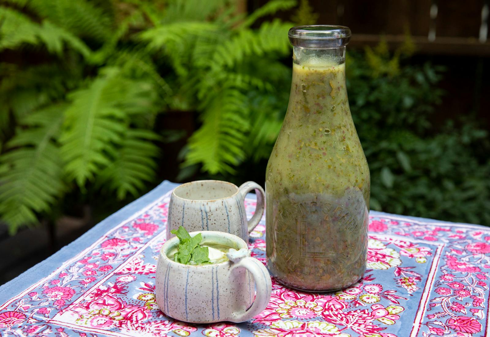 Glass bottle with schav alongside mugs of schav with sour cream and fresh sorrel atop printed tablecloth, outdoors.