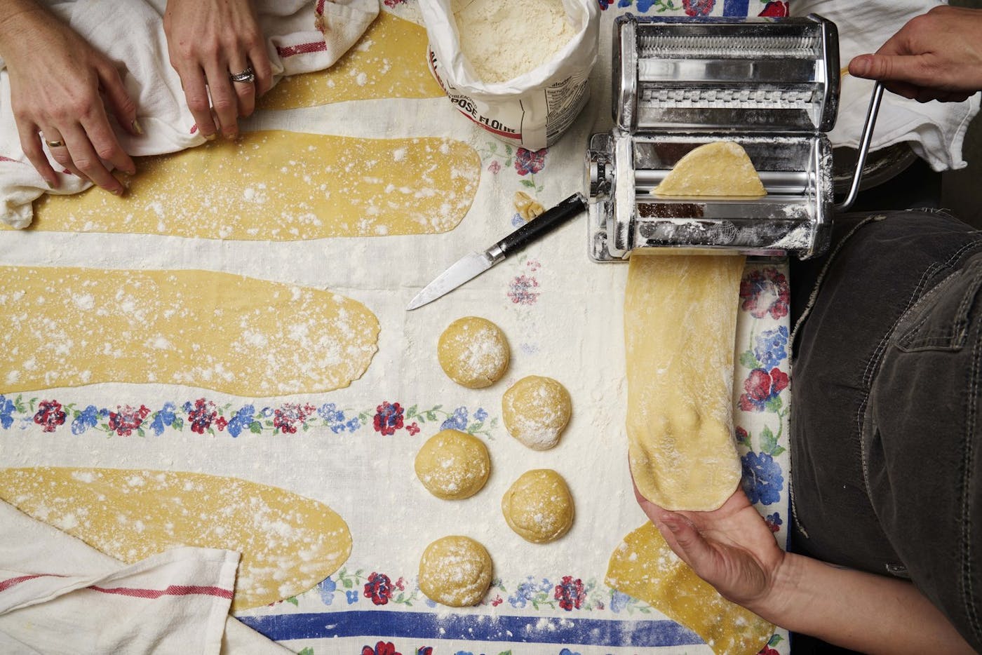 Two chefs rolling pasta dough with pasta machine atop floral tablecloth.