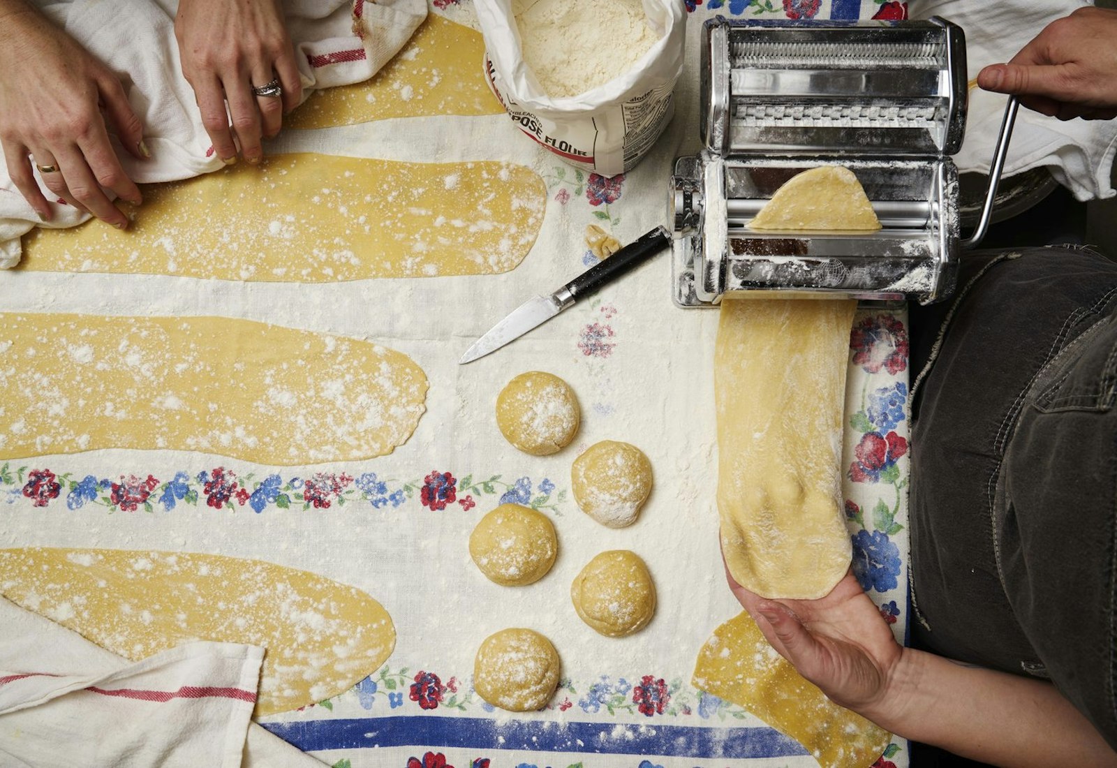 Two chefs rolling pasta dough with pasta machine atop floral tablecloth.