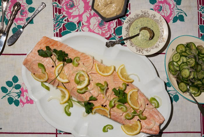 Poached salmon garnished with lemon wedges, celery and parsley alongside tonnato sauce, sauce vert and cucumber salad.