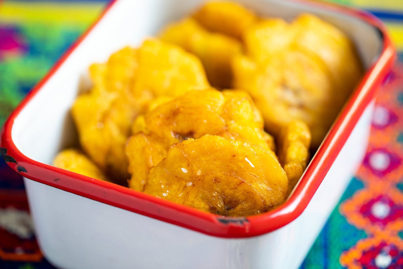 Tostones sprinkled with coarse salt in red-rimmed dish atop vibrant woven tablecloth.