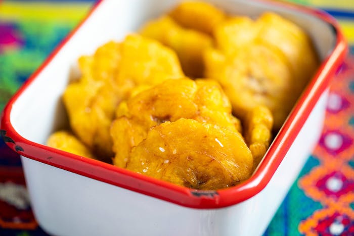 Tostones sprinkled with coarse salt in red-rimmed dish atop vibrant woven tablecloth.