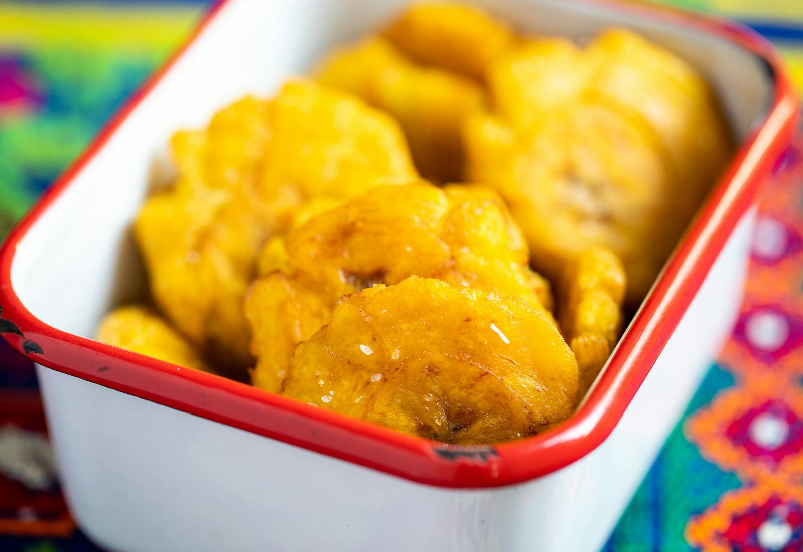 Tostones sprinkled with coarse salt in red-rimmed dish atop vibrant woven tablecloth.