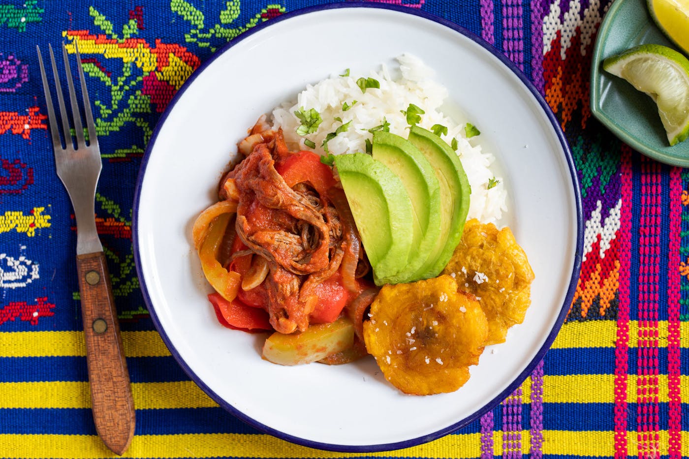 Ropa vieja with tostones, white rice and avocado on white plate alongside dish with lime wedges, atop vibrant woven tablecloth.