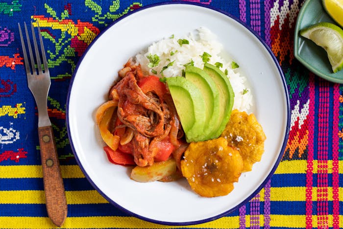 Ropa vieja with tostones, white rice and avocado on white plate alongside dish with lime wedges, atop vibrant woven tablecloth.