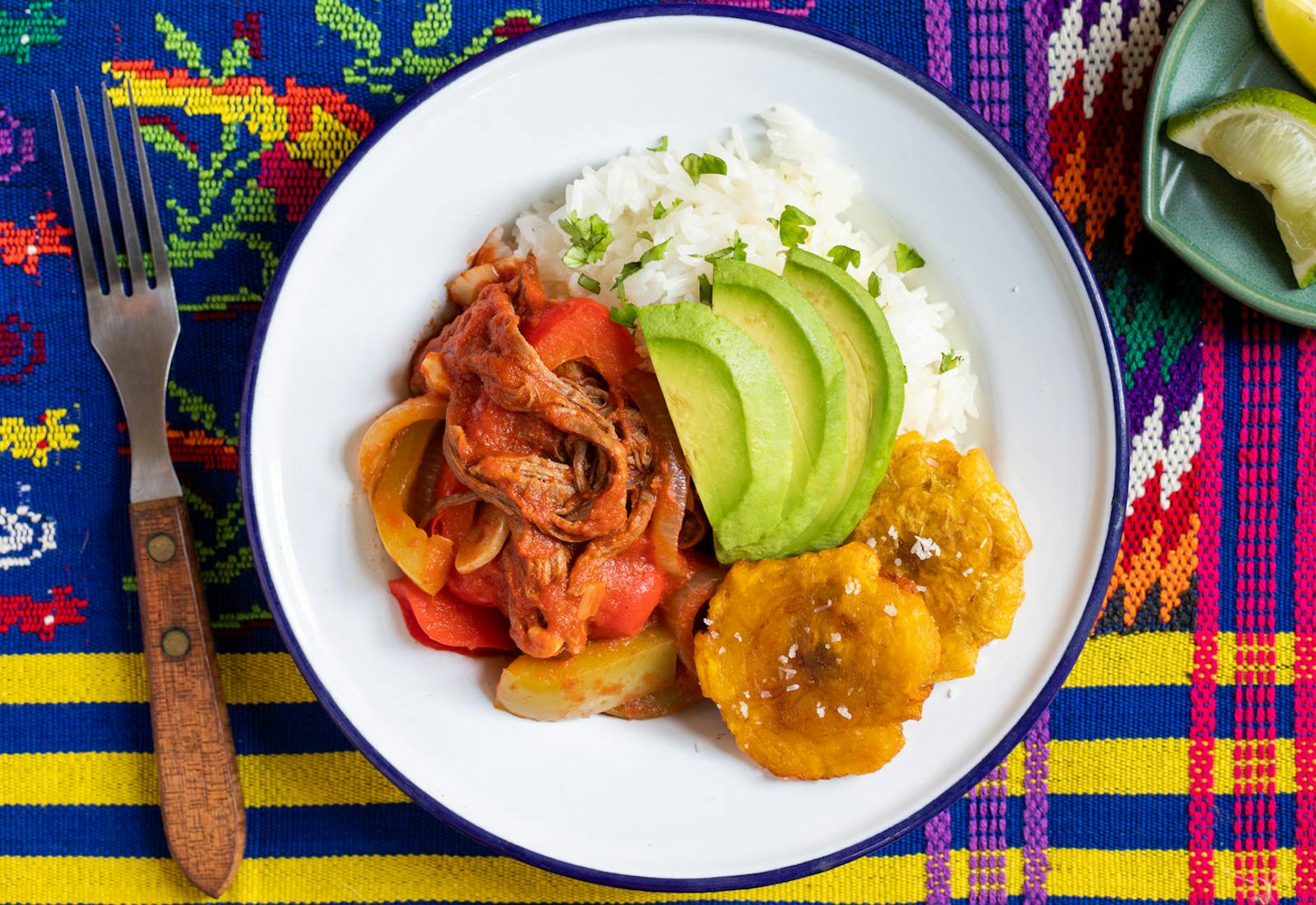 Ropa vieja with tostones, white rice and avocado on white plate alongside dish with lime wedges, atop vibrant woven tablecloth.