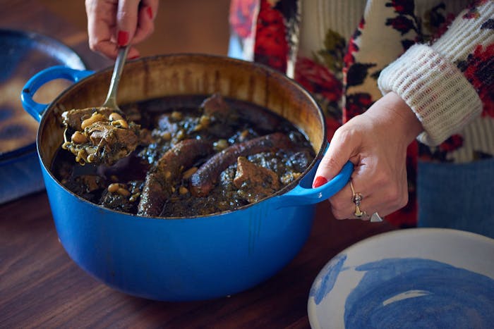 Woman serving p'karla from blue cast iron pot onto blue painted bowls atop wooden table.
