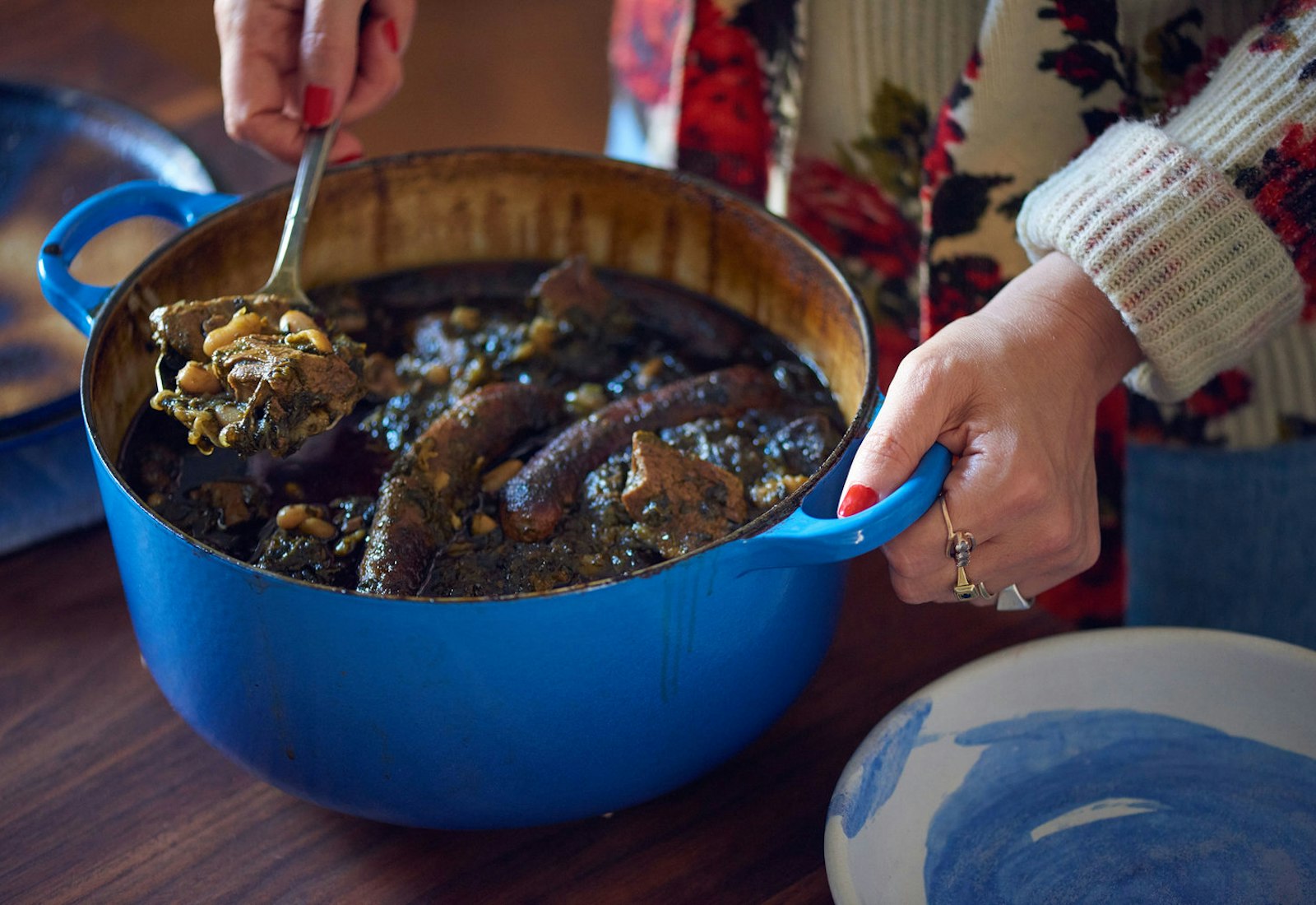 Woman serving p'karla from blue cast iron pot onto blue painted bowls atop wooden table.