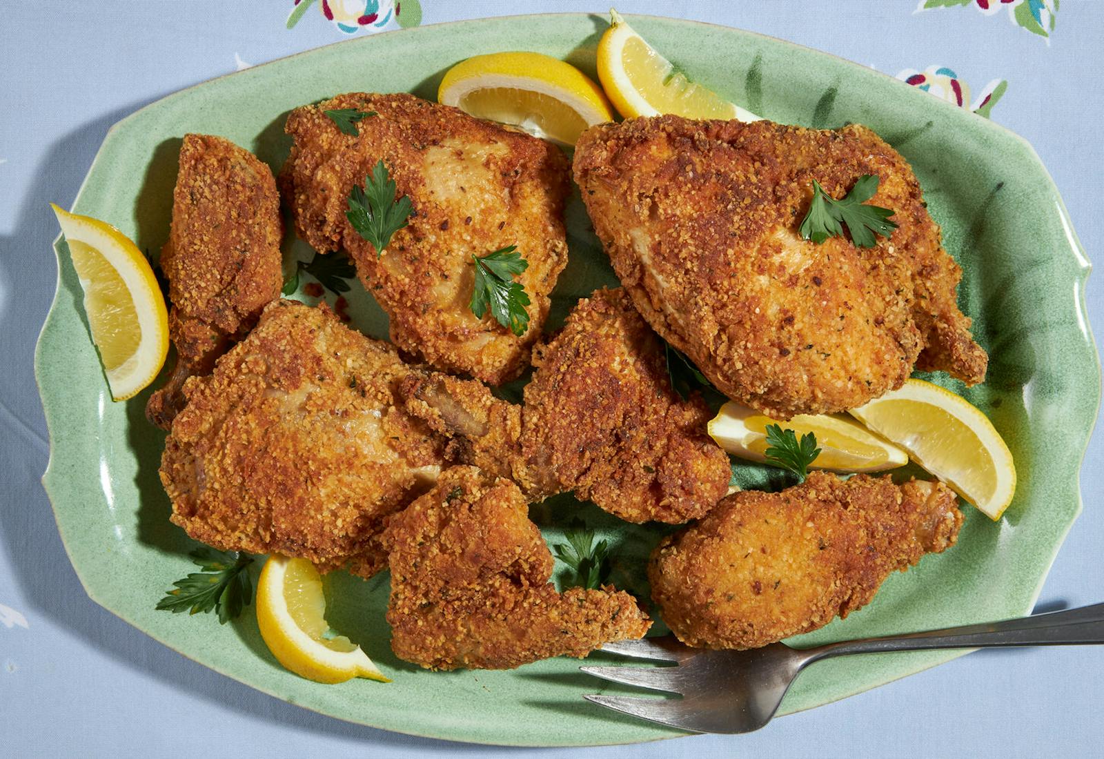 Fried chicken with parsley and lemon wedges on green platter, atop blue tablecloth. 