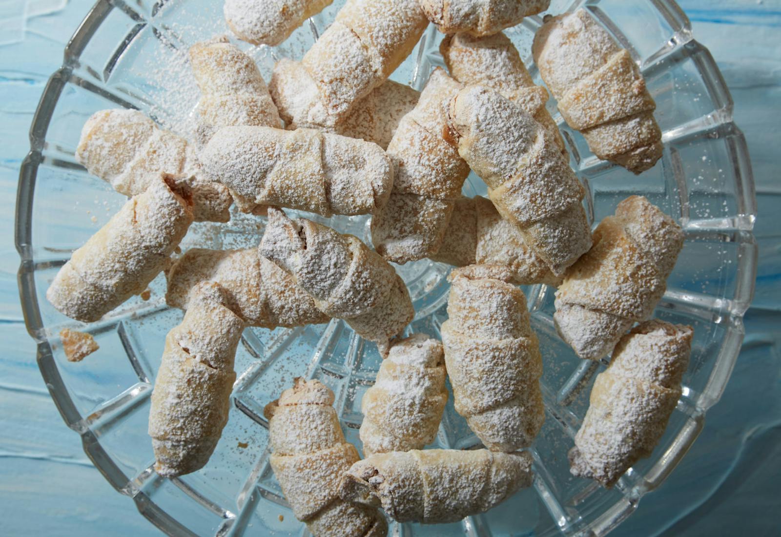 Meringue rugelach covered in powdered sugar on glass dish atop blue surface.