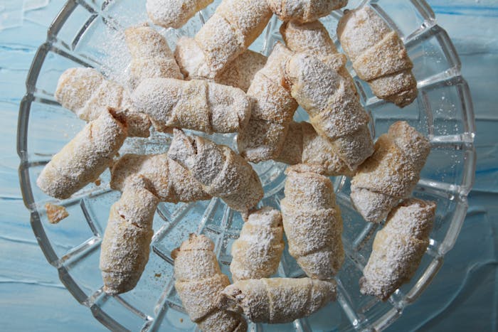 Meringue rugelach covered in powdered sugar on glass dish atop blue surface.