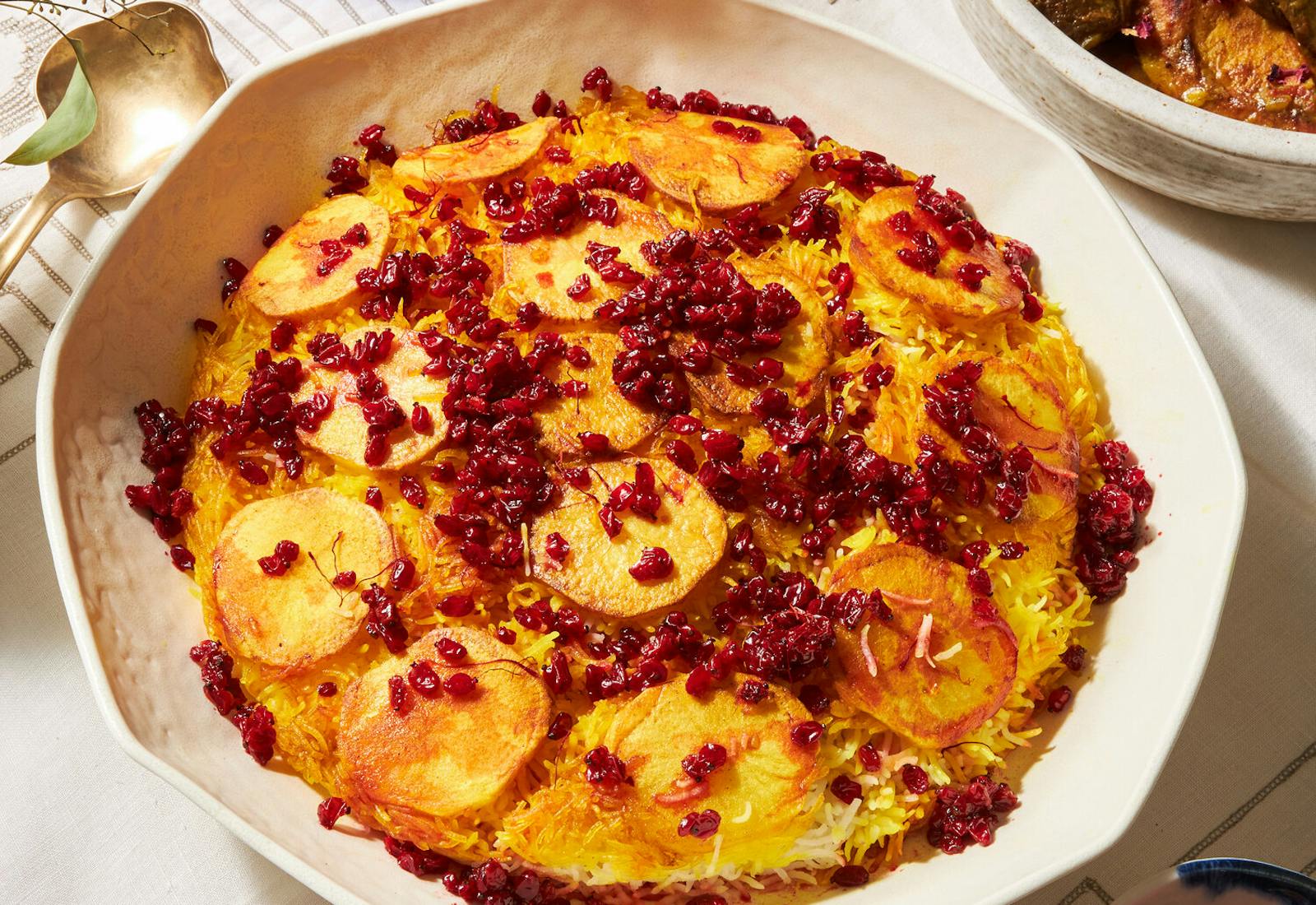 Tahdig with pomegranate kernels in large white bowl with glass of red wine and dish of rose petals.