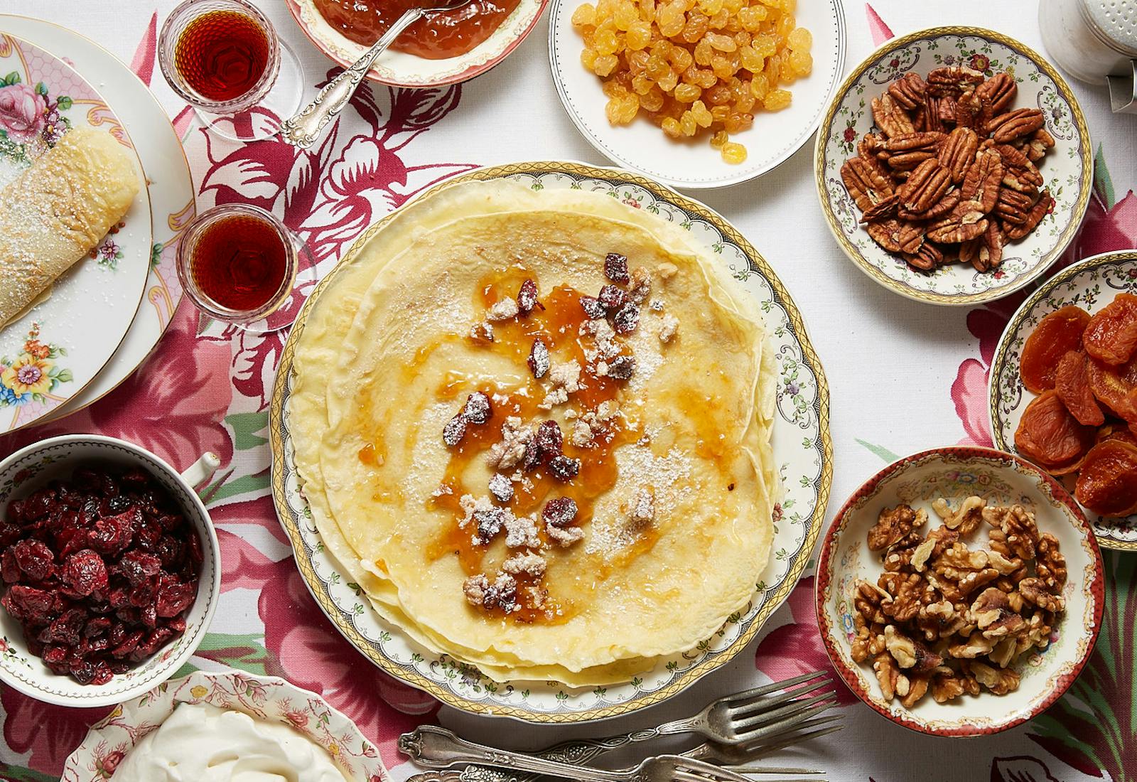 Stack of palacsinta surrounded by bowls of dried fruit, nuts, jams and sour cream, atop pink floral tablecloth.
