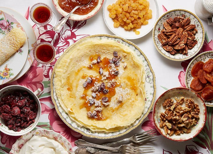 Stack of palacsinta surrounded by bowls of dried fruit, nuts, jams and sour cream, atop pink floral tablecloth.