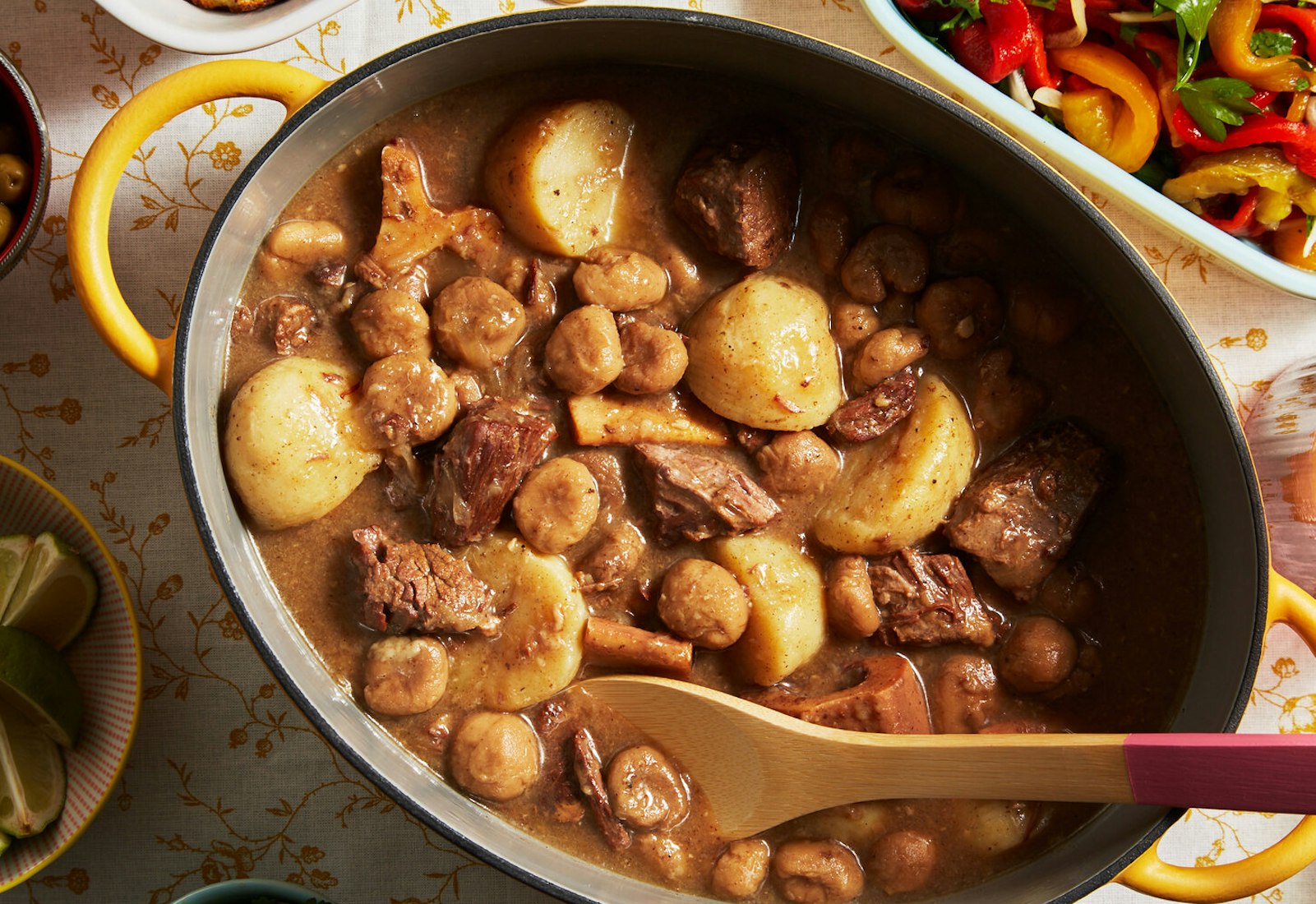 Dafina in yellow enameled cast iron dish alongside roasted pepper salad and bowl of lime wedges, atop yellow floral tablecloth.
