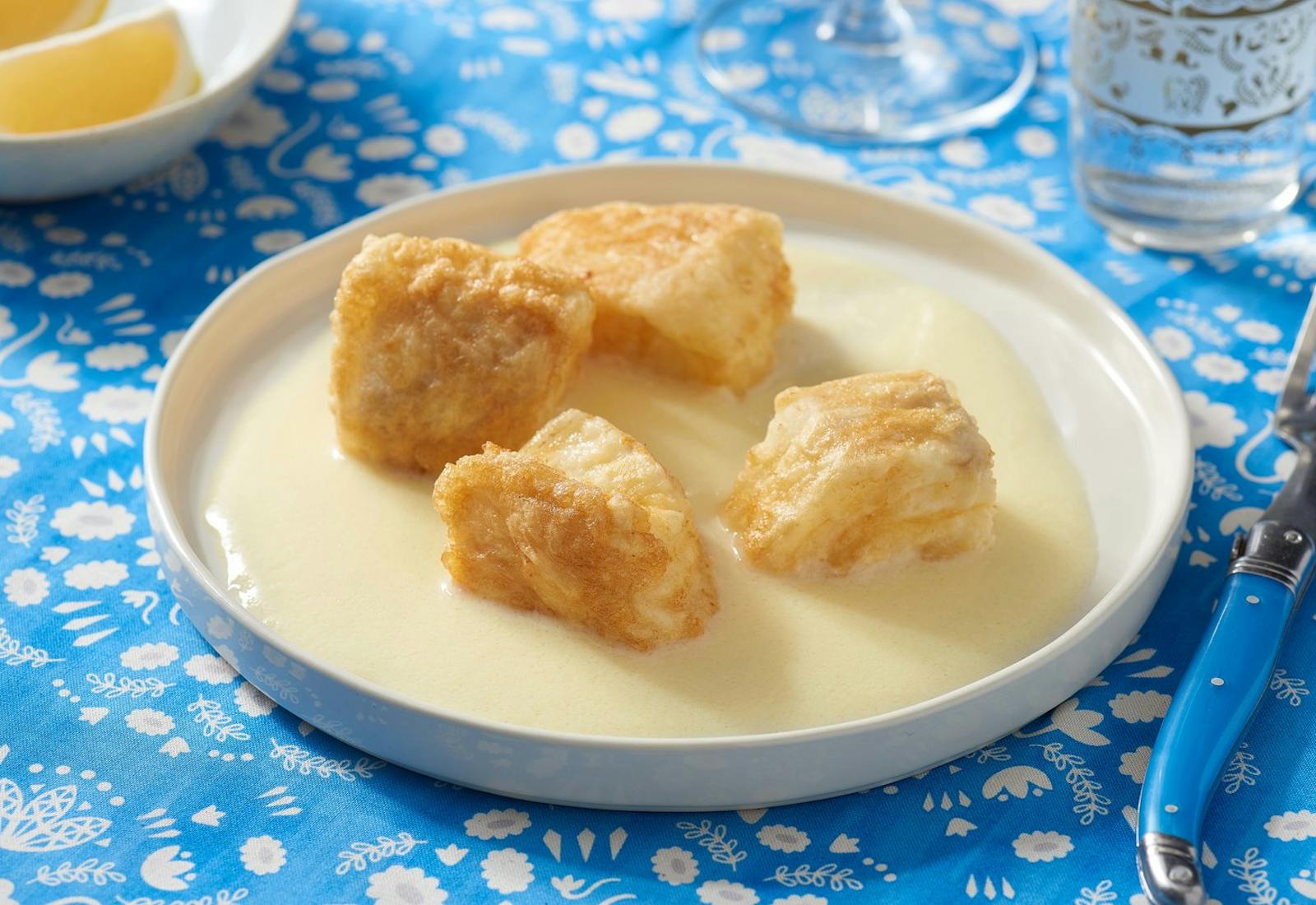 Fried with in agristada sauce in white plate alongside dish with lemon wedges atop bright blue table cloth.