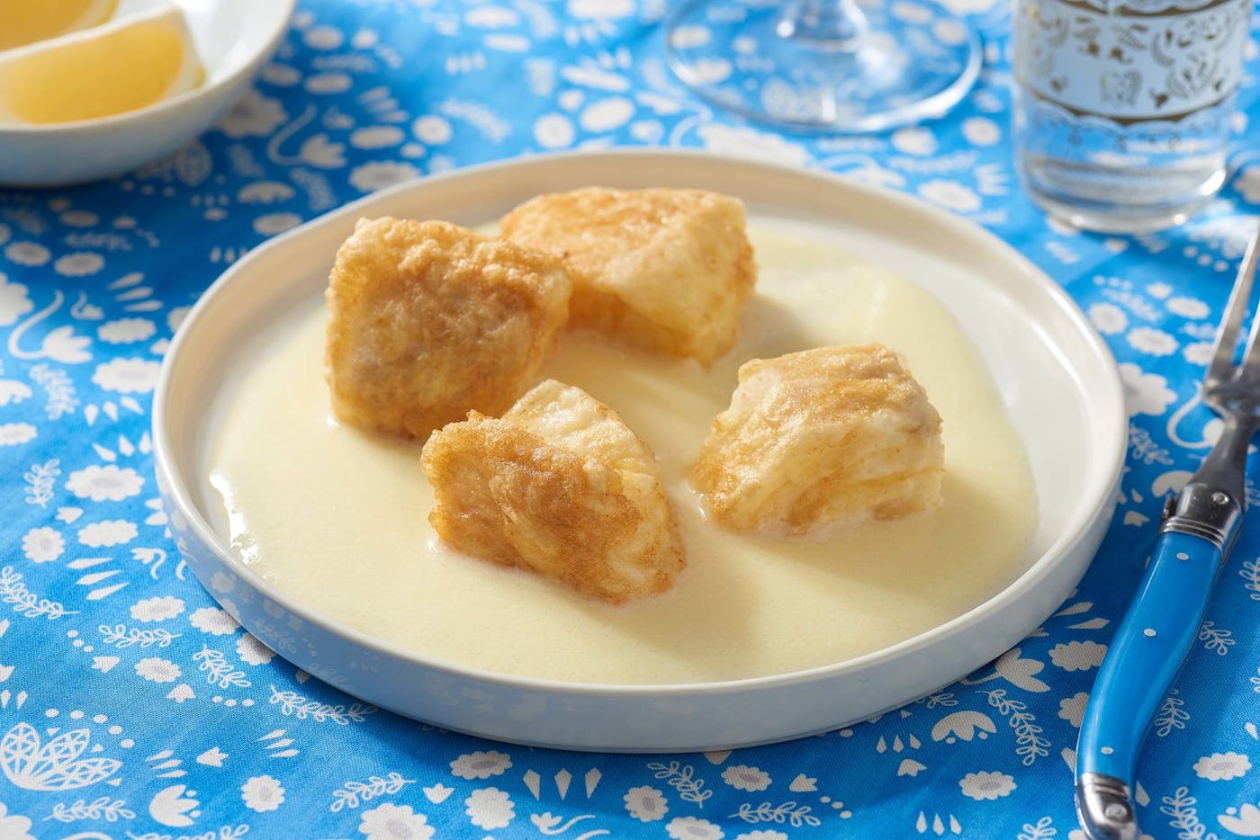 Fried with in agristada sauce in white plate alongside dish with lemon wedges atop bright blue table cloth.