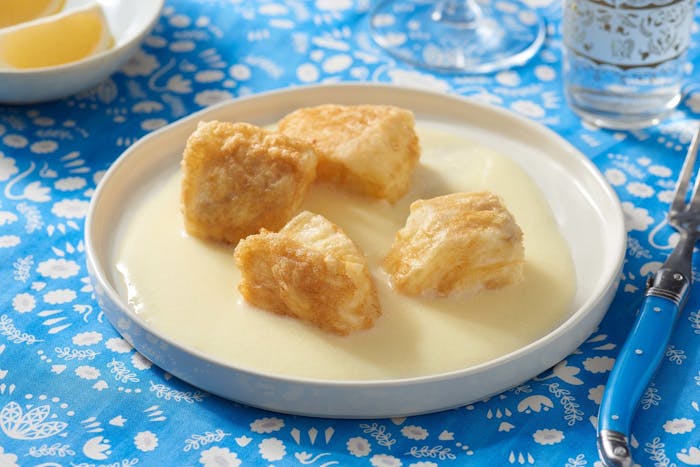 Fried with in agristada sauce in white plate alongside dish with lemon wedges atop bright blue table cloth.