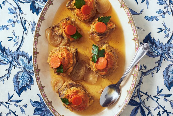 Gefilte fish in sauce with parsley, carrots and onions on oblong serving dish atop blue floral tablecloth.