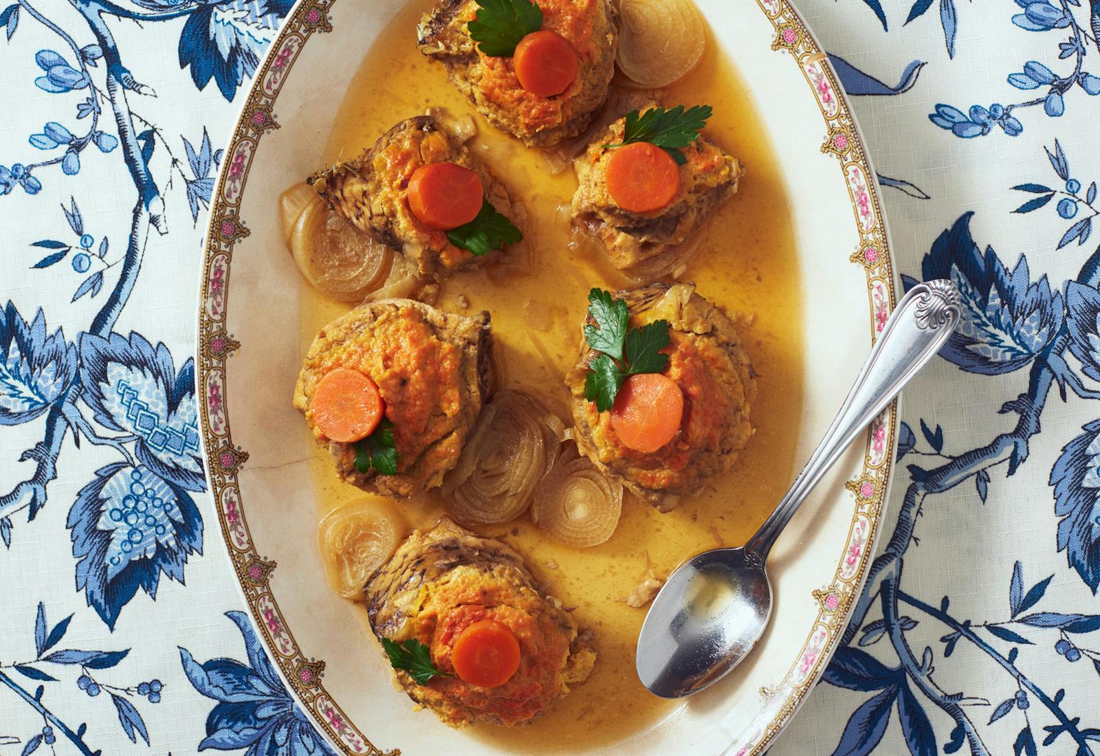 Gefilte fish in sauce with parsley, carrots and onions on oblong serving dish atop blue floral tablecloth.