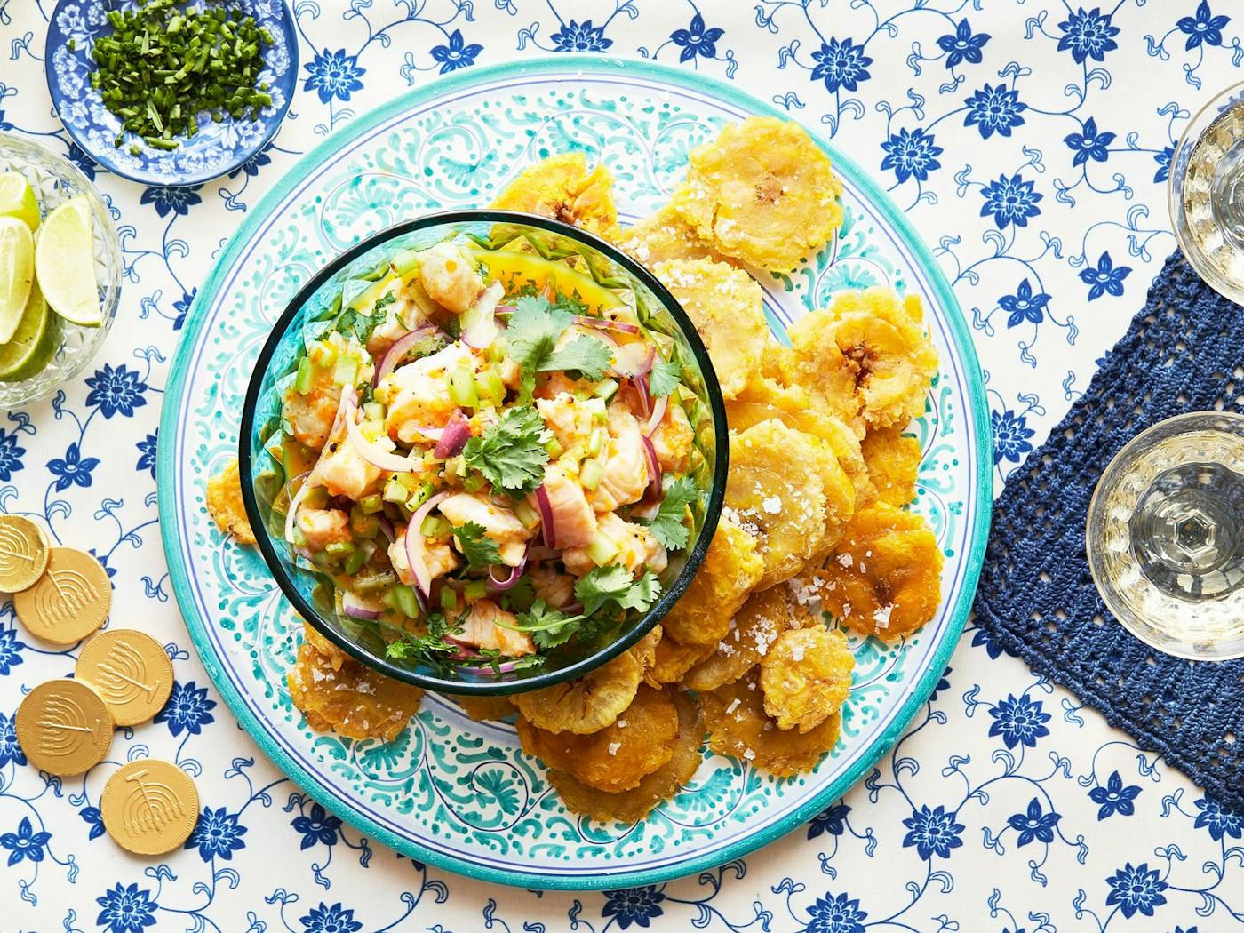 Ceviche with cilantro, tostones, limes, chocolate gelt and white wine atop blue tablecloth.