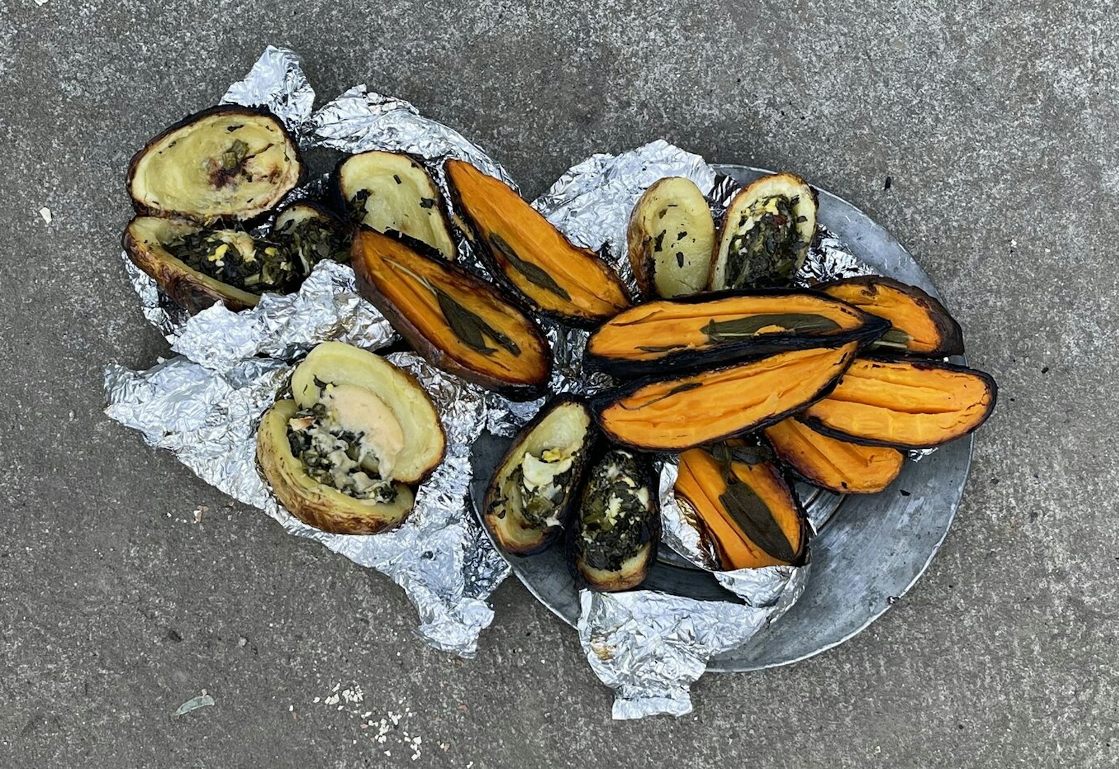 Bowl with fire-cooked sweet potatoes and stuffed white potatoes atop concrete surface.