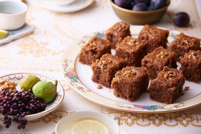 Portions of honey walnut cake on floral dish alongside plates of figs, walnuts and elderberries and mugs of lemon tea.
