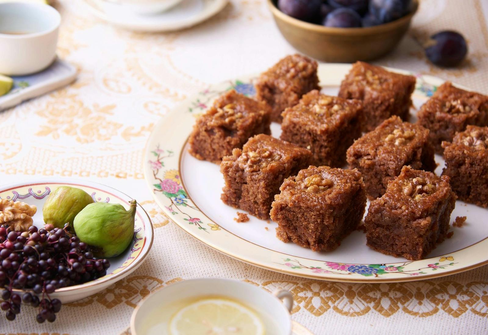 Portions of honey walnut cake on floral dish alongside plates of figs, walnuts and elderberries and mugs of lemon tea.