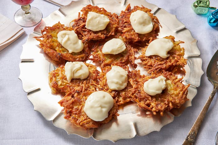 Latkes with dollops of stracchino on silver plate, red wine and dreidels atop white tablecloth.