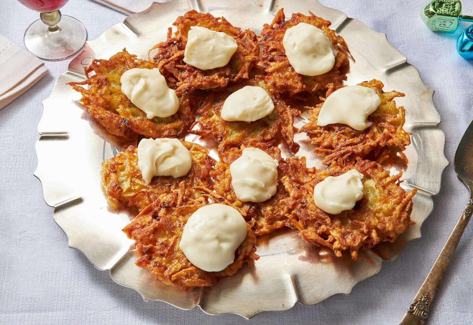 Latkes with dollops of stracchino on silver plate, red wine and dreidels atop white tablecloth.