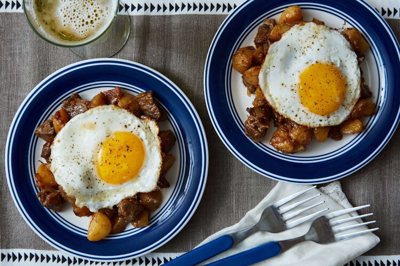 Danish hash topped with sunny side up eggs alongside glass of white wine atop grey tablecloth.