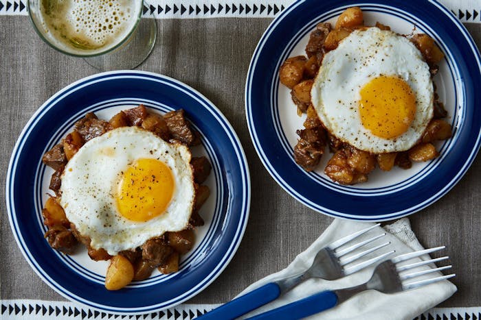 Danish hash topped with sunny side up eggs alongside glass of white wine atop grey tablecloth.