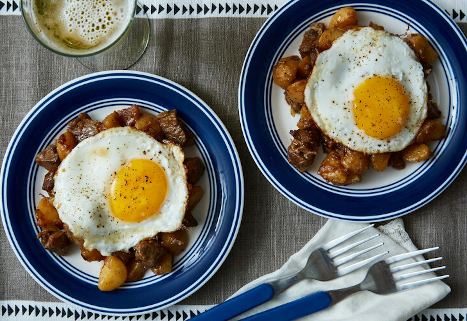 Danish hash topped with sunny side up eggs alongside glass of white wine atop grey tablecloth.