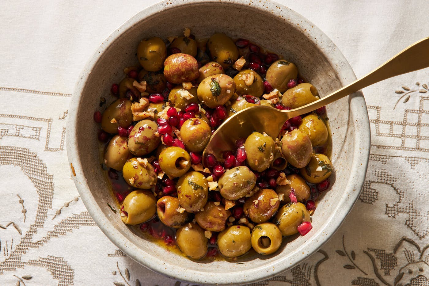 Olive salad with pomegranate in white bowl with gold serving spoon, atop white patterned tablecloth.