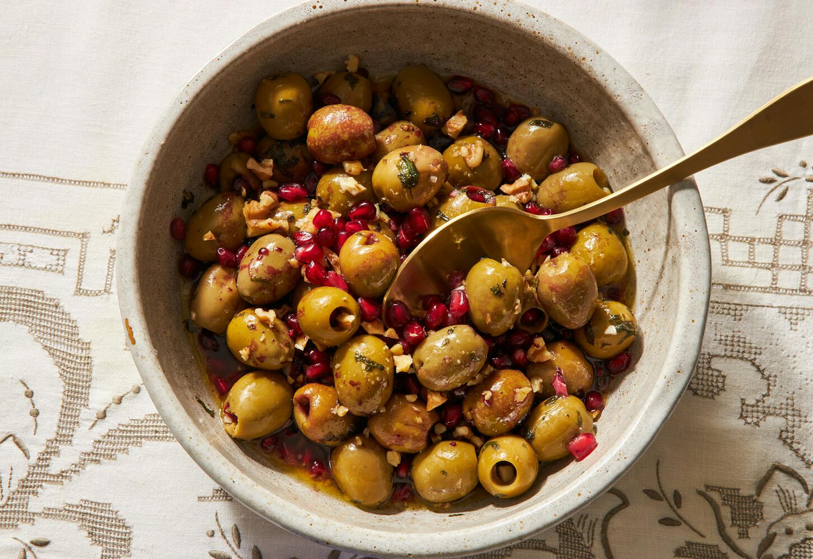 Olive salad with pomegranate in white bowl with gold serving spoon, atop white patterned tablecloth.