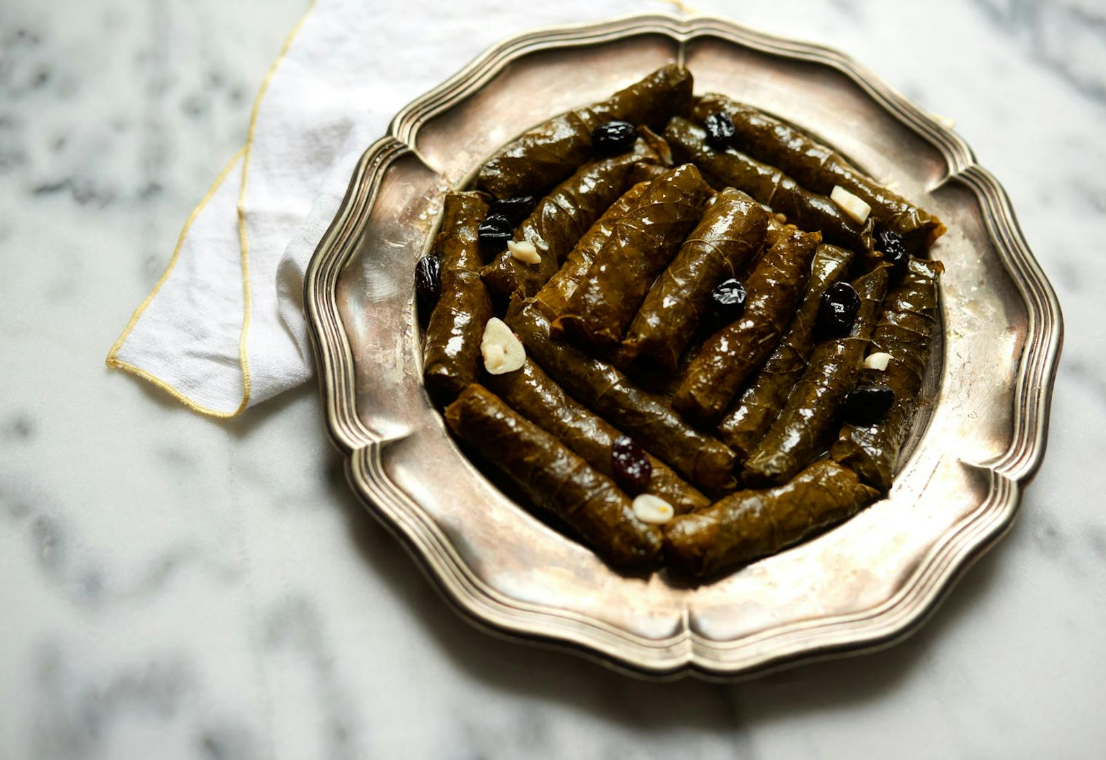 Stuffed grape leaves with slivered garlic and raisins on scalloped metal plate atop marble table.