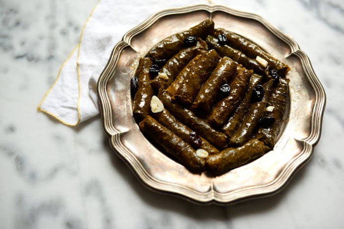 Stuffed grape leaves with slivered garlic and raisins on scalloped metal plate atop marble table.