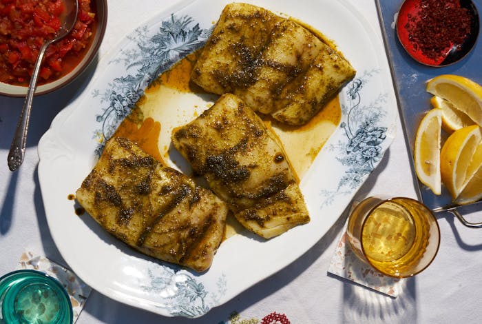 Sea bass with chermoula on grey and white plate alongside lemon wedges and bowl of chopped tomatoes.